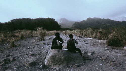 Rear view of men sitting on rock against sky