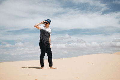 Full length of young man standing on sand against cloudy sky