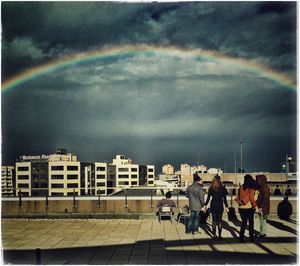 People on rainbow in city against cloudy sky