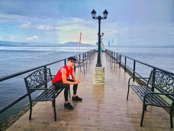 Man sitting on railing by sea against sky