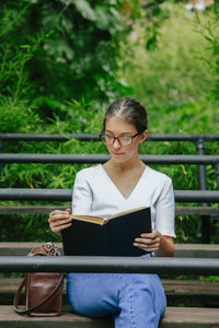 Young woman wearing eyeglasses reading book while sitting on bench in park