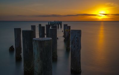 Wooden posts in sea against orange sky during sunset