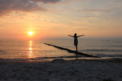 Silhouette woman with arms outstretched standing on wooden posts in sea against orange sky