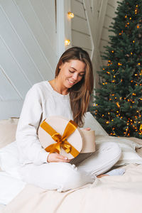 Young woman with present gift box on bed in room with christmas tree at home, happy new year