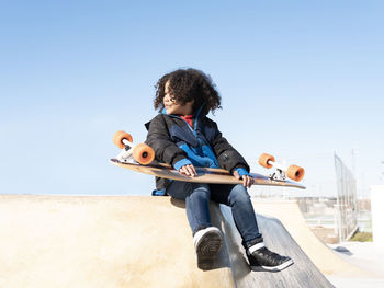Ethnic child with curly hair sitting on ramp with longboard in skate park and looking away