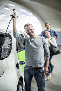 Portrait of happy man holding electrical charger with friends standing in background at gas station
