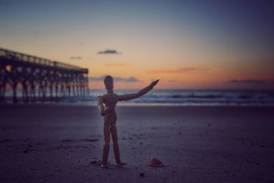 Figurine standing at beach against sky during sunset