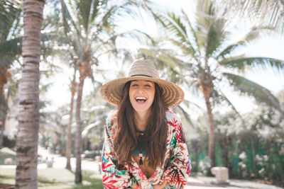 Portrait of cheerful woman wearing hat standing against palm trees