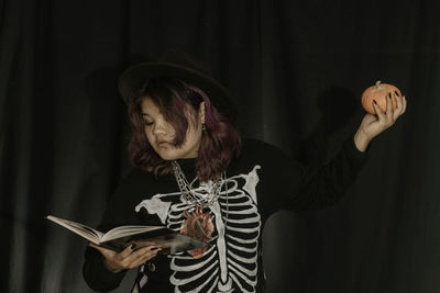 Young asian teen woman in costume witch hold pumpkin and book on black cloth background of halloween