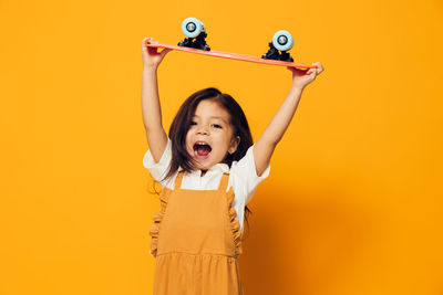 Portrait of girl holding skateboard standing with arms raised against orange background