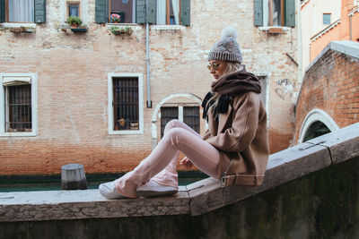 Side view of young woman sitting on retaining wall against building