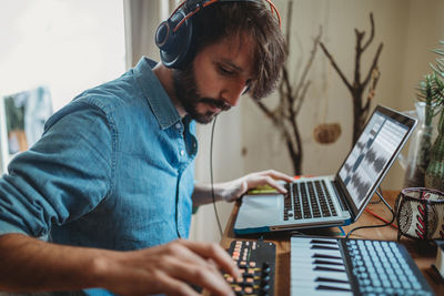 Side view of young man in headphones using synthesizer and laptop at table at home
