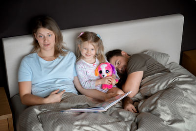 High angle view of mother reading book for daughter while father sleeping on bed at home