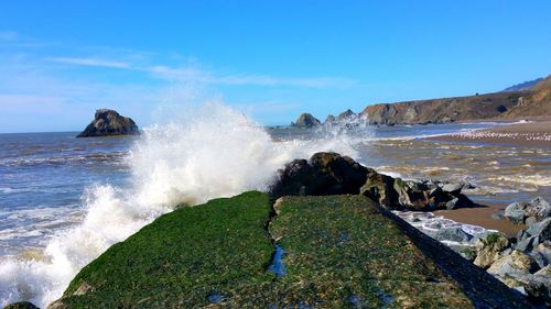 Scenic view of waves crashing on jutting breakwall in sea against blue sky