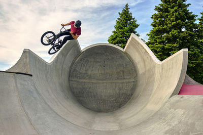 Male rider with bmx bike jumping on concrete ramp against sky at skateboard park