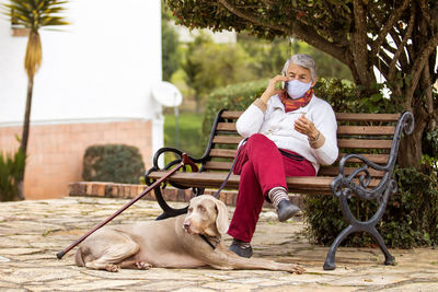 Full length of senior wearing mask with dog sitting outdoors