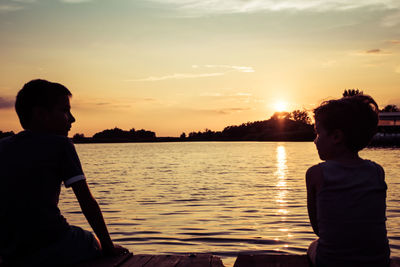 Rear view of boys sitting by lake during sunset