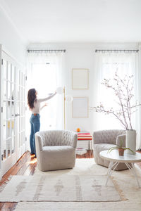 Woman adjusting curtains in white, bright room w/ modern furniture