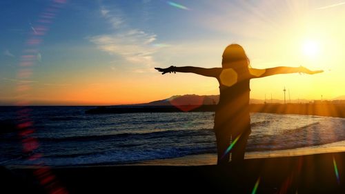 Rear view of woman with arms outstretched while standing on beach during sunset