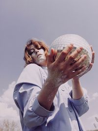 Low angle portrait of woman with glitter on face holding disco ball against clear sky