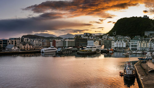 Sailboats moored on river by buildings against sky during sunset