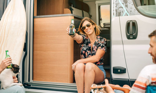 Woman showing beer bottle having fun with friends sitting at the door of their camper van