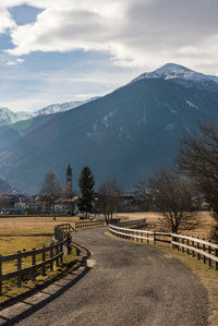 Pathway between trees leading to alpine village. cloudy sky and snow-capped mountains italian alps.