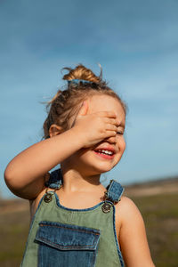 Delighted adorable little girl in overalls standing with hand on face in meadow and closed eyes