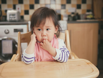 Baby girl with fingers in mouth sitting on high chair at home