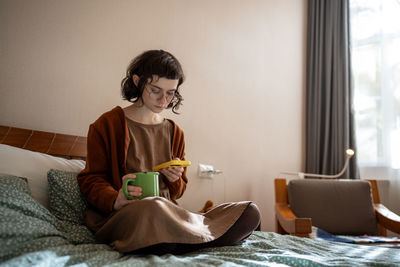 Young woman reading book while sitting on bed at home