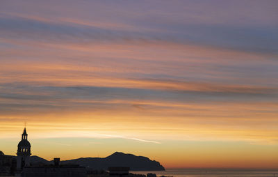 Silhouette buildings by sea against orange sky