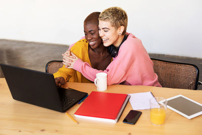 Cheerful lesbian couple embracing while working over laptop on table at home