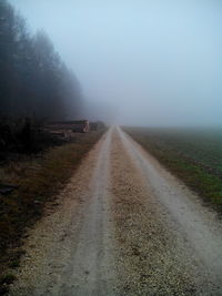 Dirt road amidst trees against sky during foggy weather