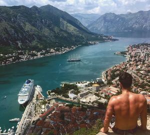 Rear view of shirtless man sitting on rock against town by sea