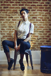 Portrait of young man wearing suspenders sitting on chair against brick wall