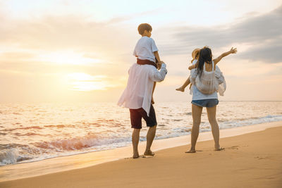 Rear view of couple standing at beach against sky during sunset