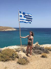 Full length of young woman holding greek flag while standing at beach against clear sky