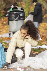 Young female volunteer talking on smart phone while collecting garbage in park