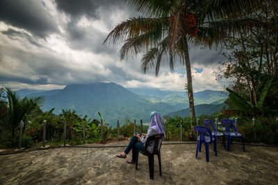 Rear view of woman sitting on chair by palm trees