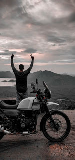 Rear view of man with arms raised standing by motorcycle against cloudy sky