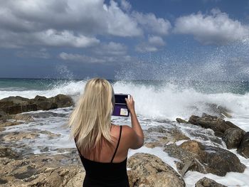 Rear view of woman photographing sea with mobile phone at beach against sky