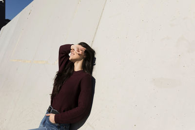 Woman with hands in pockets shielding eyes while leaning on wall during sunny day