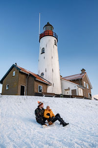 Full length of couple sitting on snow covered land against lighthouse