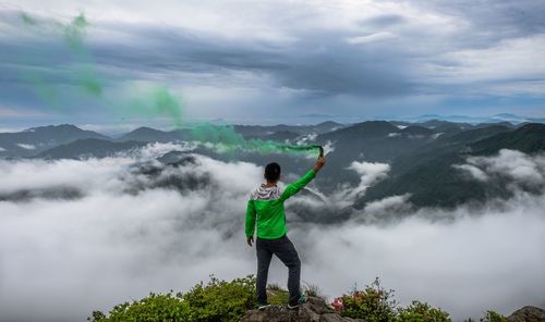 Rear view of man holding distress flare on mountain against cloudy sky