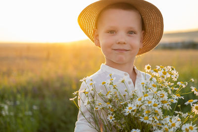Portrait of boy wearing hat holding flowers on field