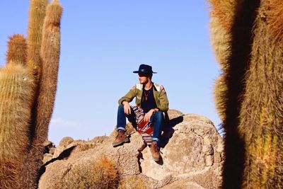 Man sitting on rock by cactus against clear sky