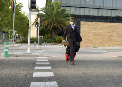 Serious black male entrepreneur wearing medical mask crossing road in city while walking with bags towards airport during coronavirus epidemic