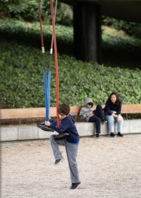 Boy playing with colourful steel swing ropes. brother and mother sitting watching in the background.