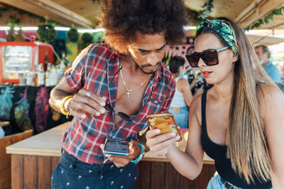 African american man showing photo on mobile phone to girlfriend while standing near counter of outdoor beach bar