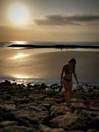 Woman standing on rock at beach against sky during sunset